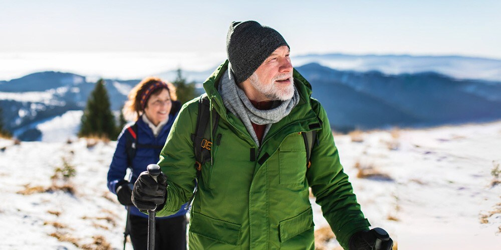 A man and woman hiking in a snowy landscape. The man wears a green jacket and knit hat, holding trekking poles, with the woman following in a blue jacket. Snowy hills and trees are in the background.