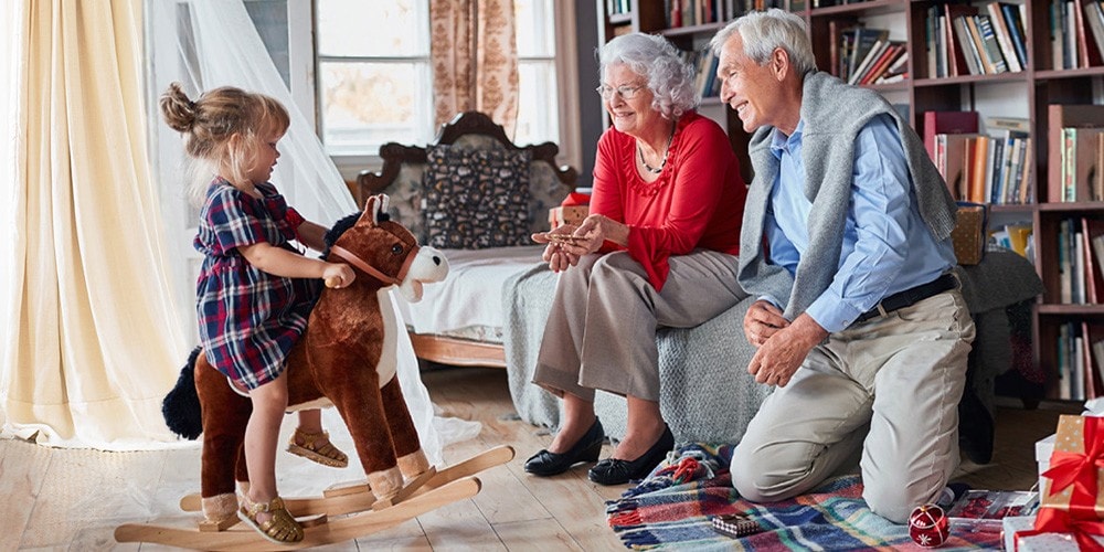 A child plays with a rocking horse, while two elderly adults smile while watching. They are sitting in a room with natural light streaming in through a window, and there are bookshelves in the background.